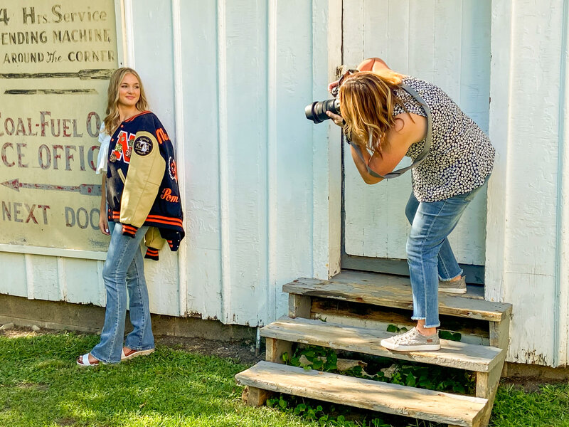female photographer  taking a picture of a high school senior girl wearing jeans with a varsity jacket draped over one shoulder.  The girl is standing against a white wood building.