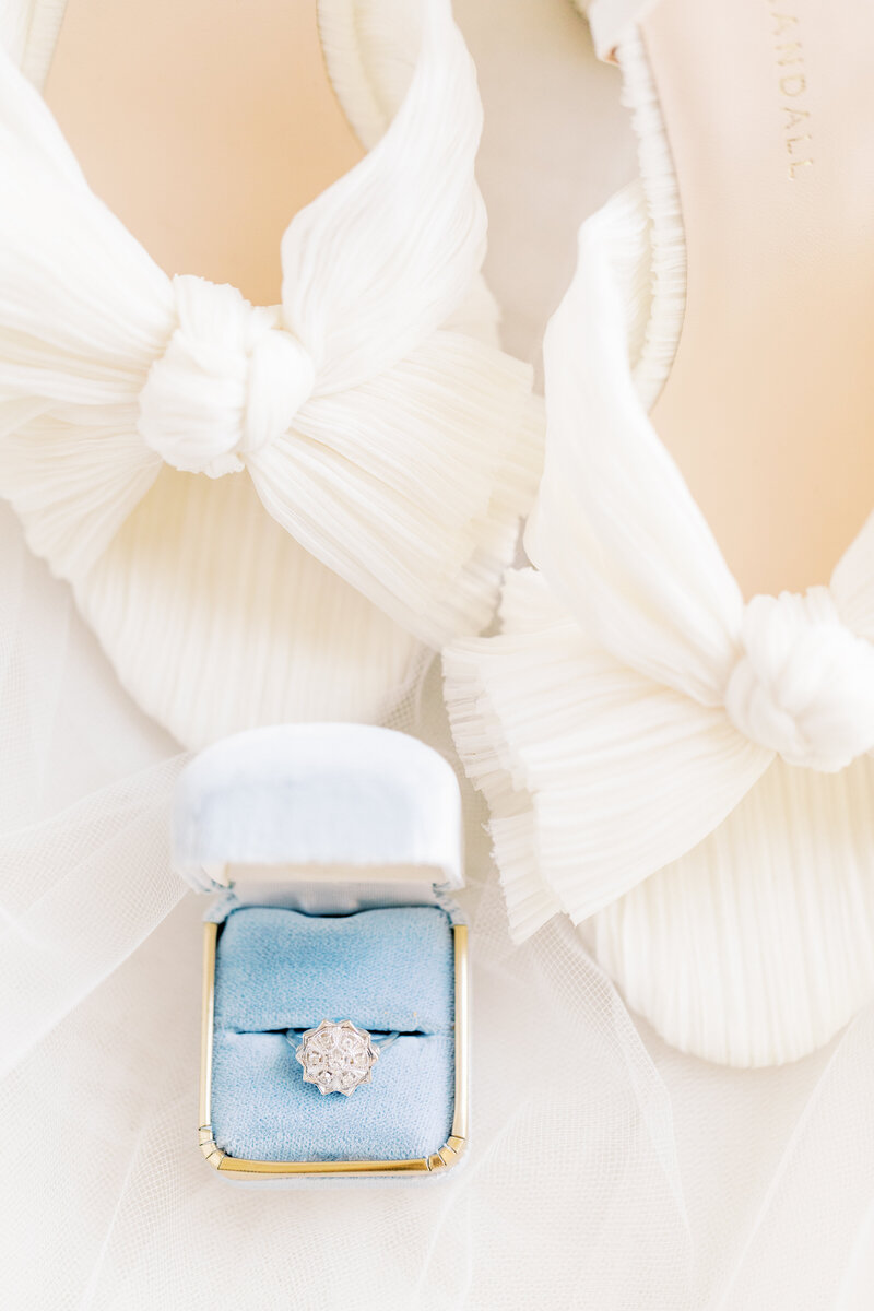 A vintage-inspired diamond ring nestled in a soft blue velvet box, set against delicate white bridal heels with elegant pleated bows