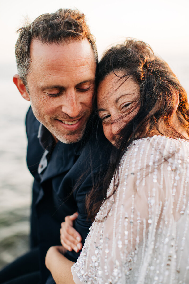 bride and groom snuggle together by the san diego ocean while the wind sweeps her hair across her face