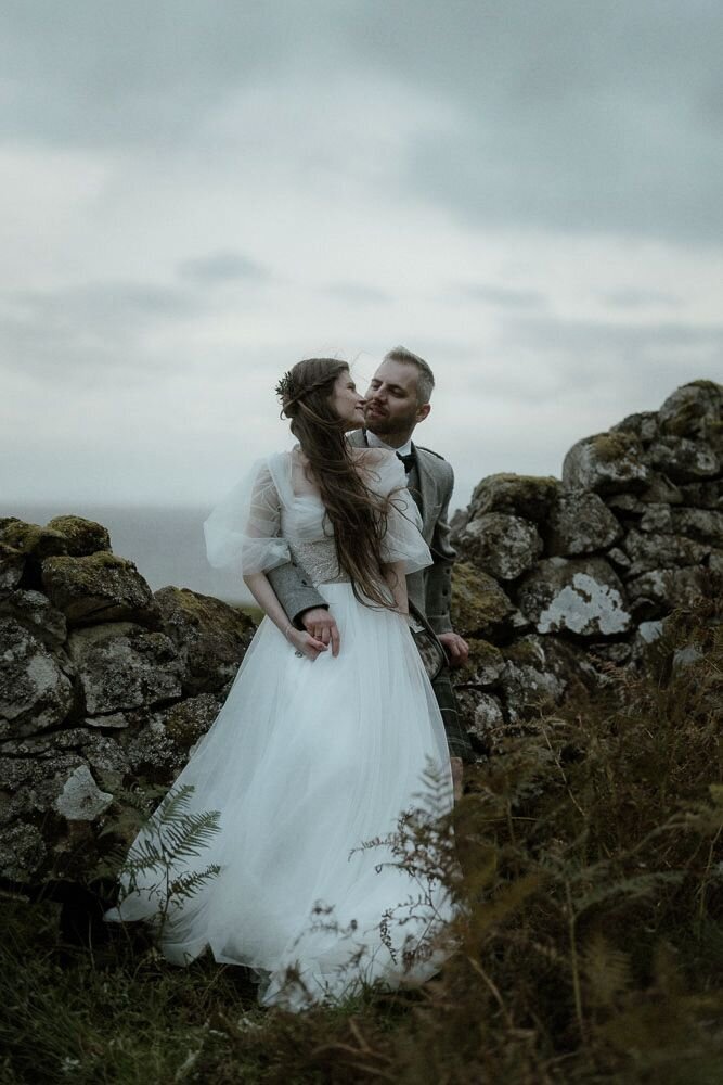 Bride and groom cuddle and hold hands while leaning against a wall on the Isle of Skye, Scotland