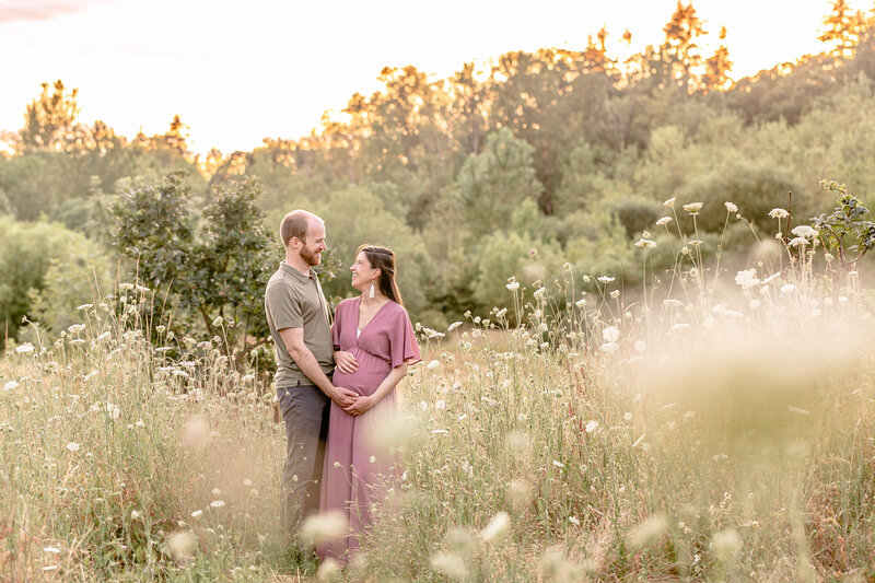 pregnant woman in mauve dress holding belly and looking at partner who is wearing an olive green shirt and grey slacks. The couple is smiling at each other and the dad also has one hand on moms belly. They are surrounded by tall, white wildflowers in a lush field at sunset.