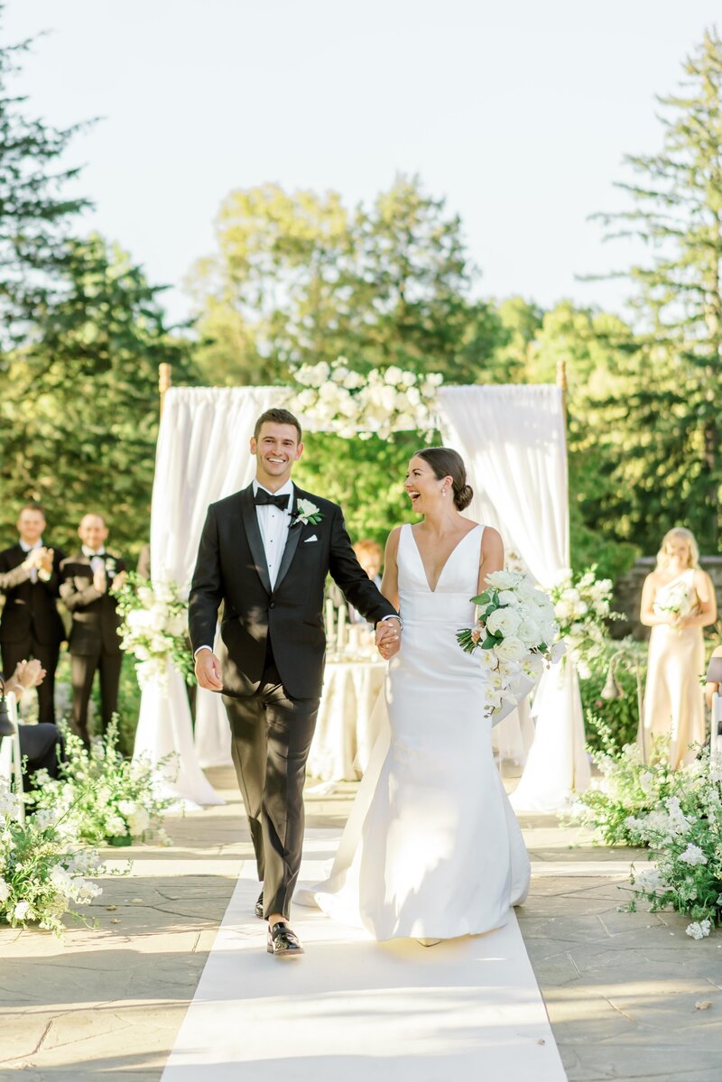 Bride and groom walking down the aisle outdoors, smiling and holding hands.