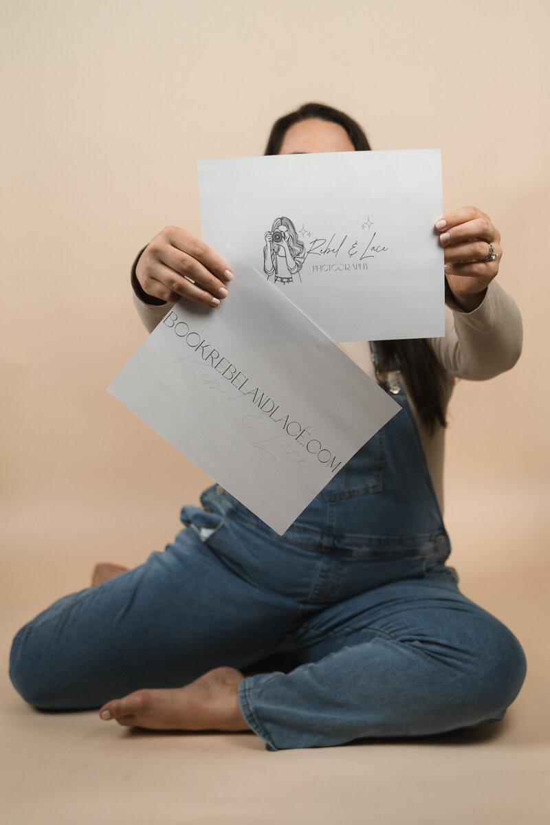 Woman holding two signs, one is the logo for Rebel and Lace, the other sign is the website address.
