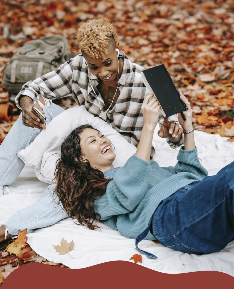 Two feminine-presenting people relax on a textured picnic blanket, laid out on crisp fall leaves. One partner lies in the lap of the other, holding up a book. They both smile widely, as they look at the book together.