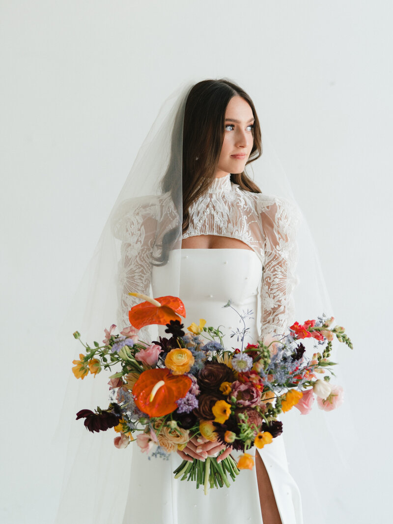 Portrait of bride with long brown hair and lace long sleeves holding a large colorful bouquet standing in front of a white studio background