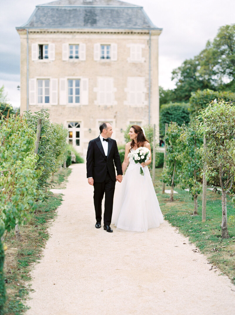 Bride and groom hold hands and walk down vineyard path outside a chateau