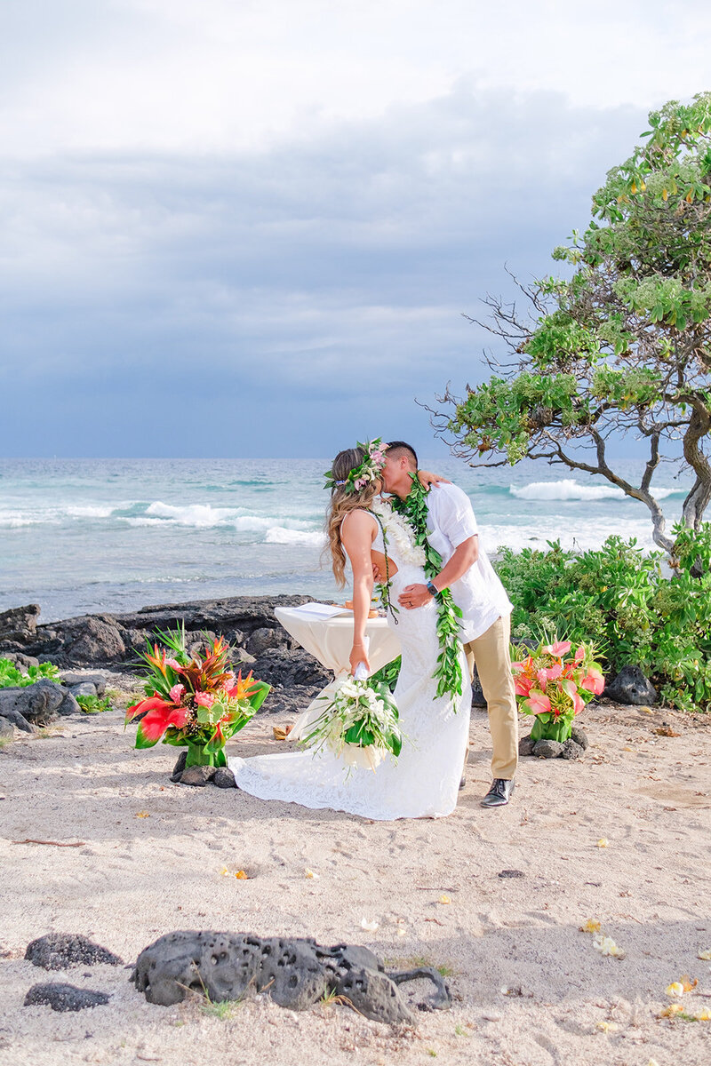bride and groom kissing on the beach with lei and bouquet