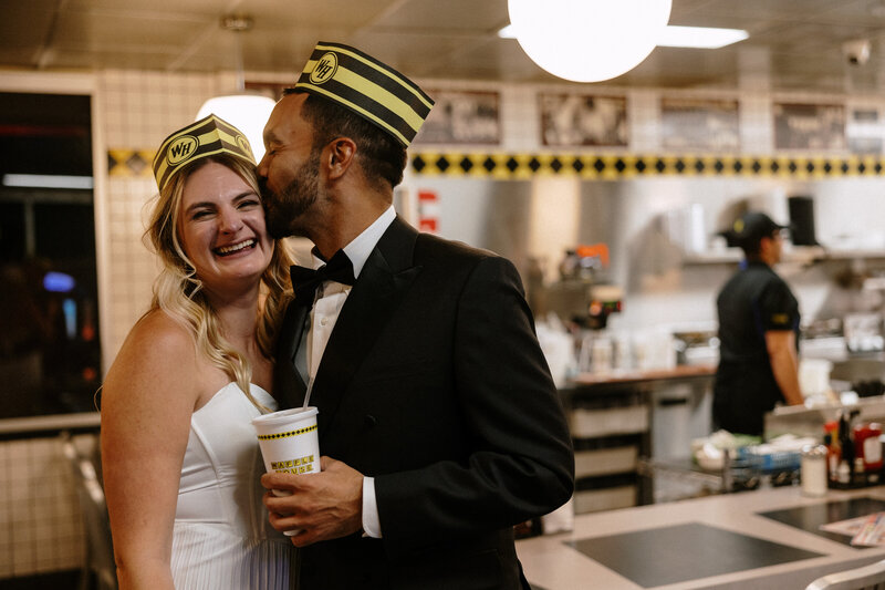 A couple in formal attire shares a moment at a diner, both wearing playful yellow and black hats, holding a drink.