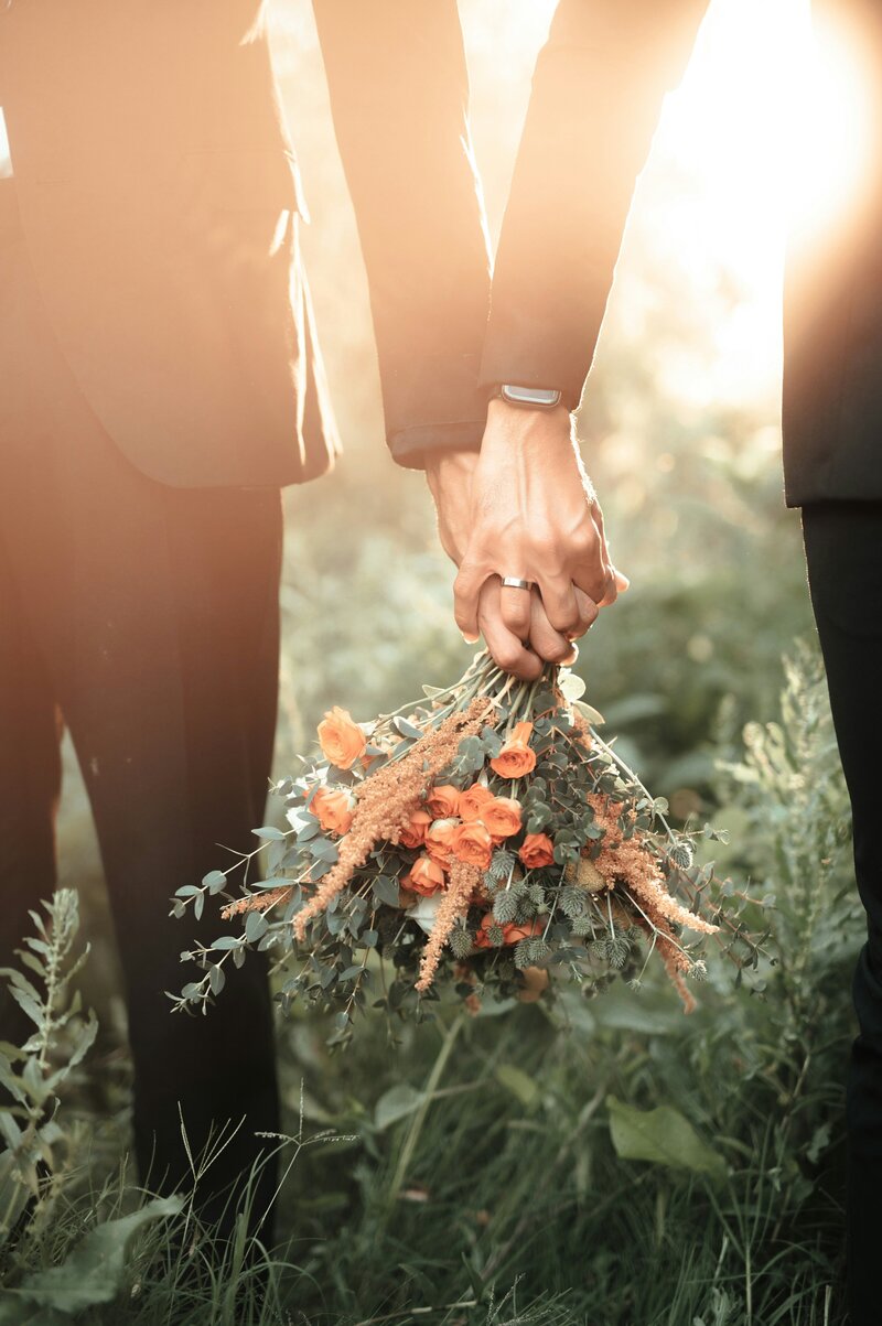 Two grooms hold hands while one carries a rustic bouquet of orange and peach flowers with greenery, set in a sunlit, grassy outdoor area.