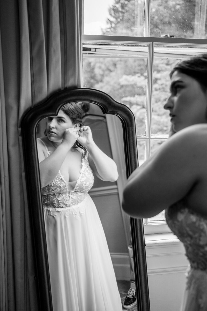 black and white image of a curvy white bride in a spaghetti-strap wedding gown looking in the mirror putting on her wedding earrings