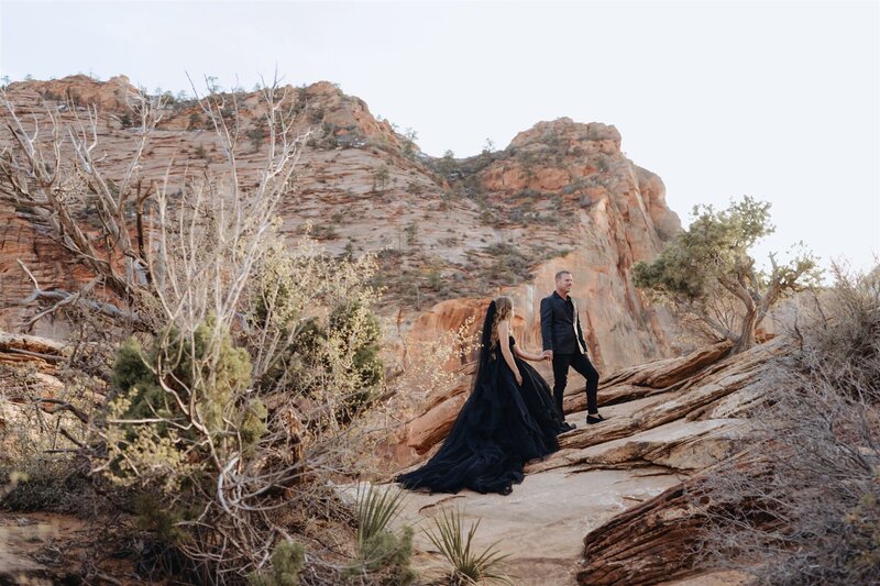 Bride and groom both dressed in black hiking up to their ceremony during their elopement in Zion.