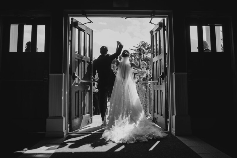 Black and white photo of Alexa Nahas photographing the bride and groom exiting the church, camera close to her face.