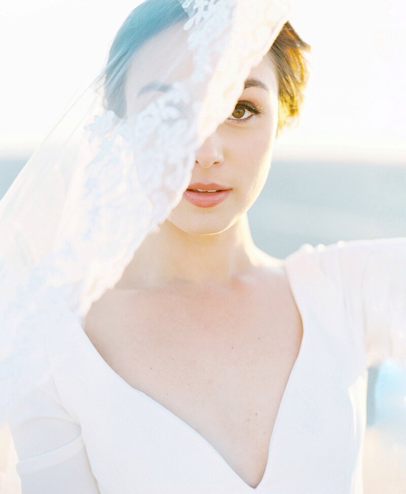 Kohnur bride with natural, soft makeup and elegant updo behind a lace veil, wearing a long-sleeved gown at