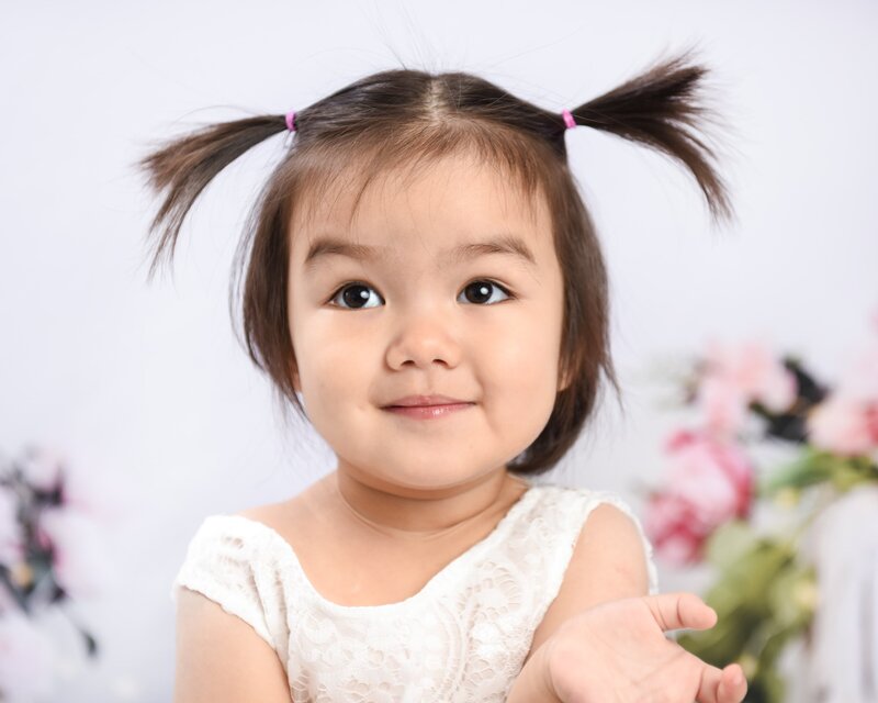 A toddler girl with pigtails wearing a white lace dress, smiling sweetly with her hands raised, in a soft floral background setting.