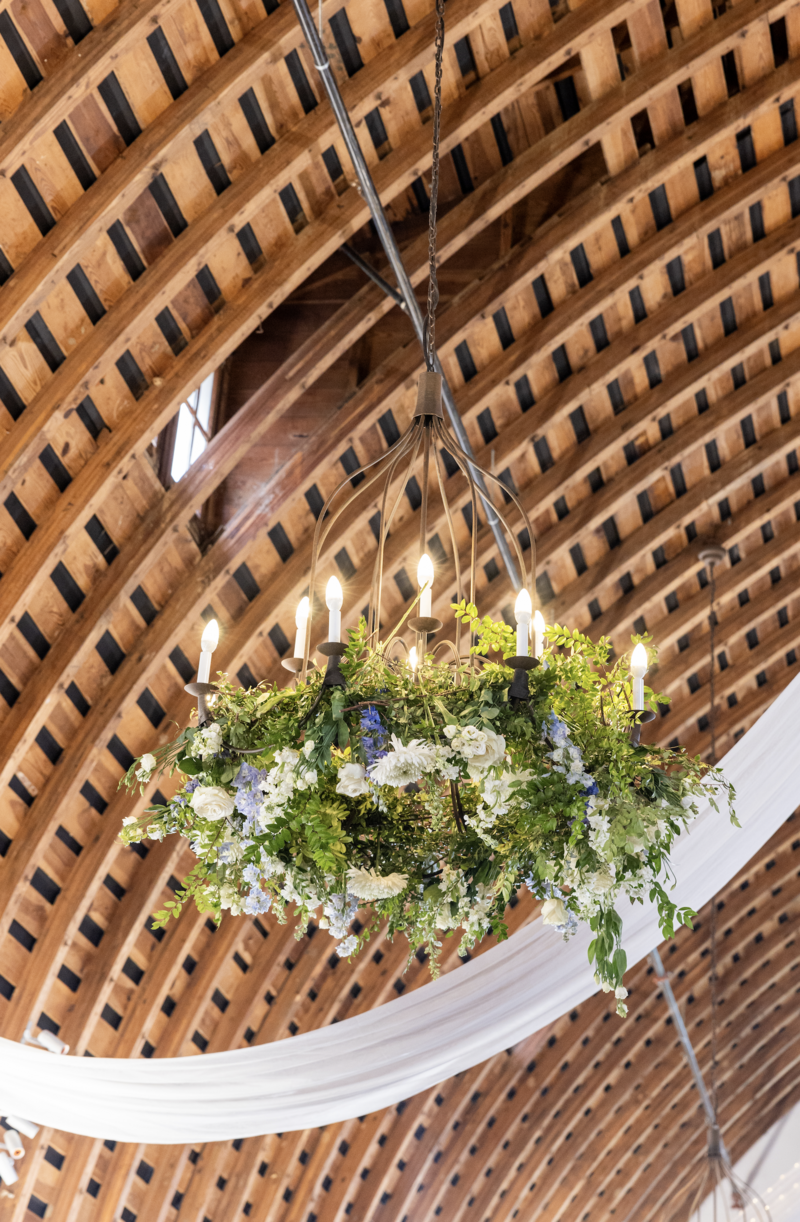 Spring wedding at WinMock at Kinderton, designed by Charming Carolina — close-up of chandelier decorated with lush greenery and dusty blue floral accents, hanging under soft white draping and fairy lights.