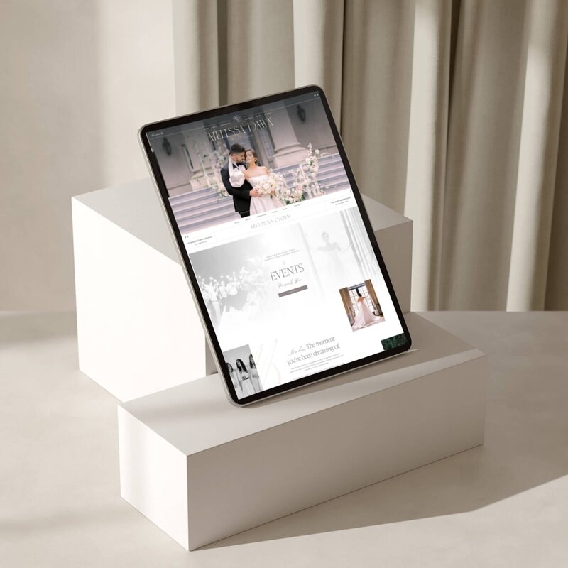 A tablet displaying a wedding planning website is elegantly placed on white geometric stands, set against a beige curtain background. Perfect for showcasing our branding and website design packages tailored specifically for creatives looking to make their mark in the industry.