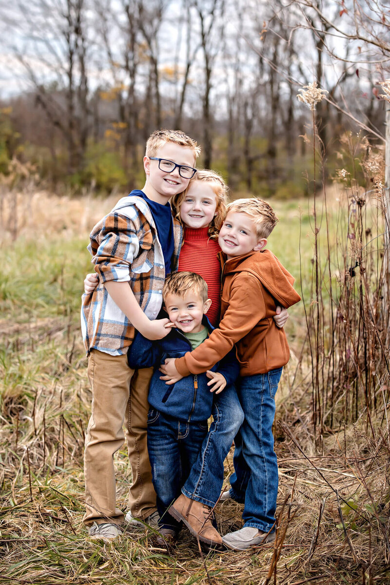 Four young children standing in a grassy field in the fall foliage hugging each other and smiling at the camera.