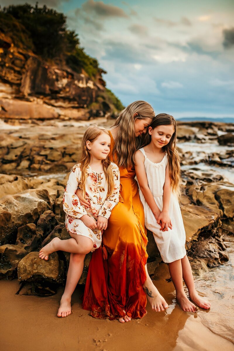 Mother with 2 young daughters cuddling on rocks near the beach