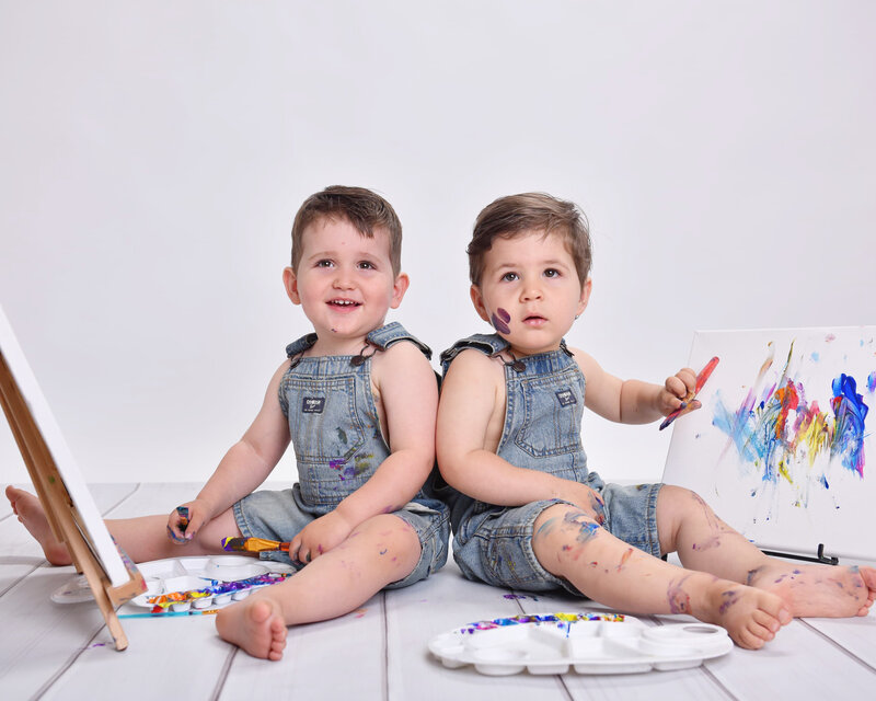 Twin toddlers in matching blue overalls painting on a canvas together during a playful toddler photo shoot with an art theme.