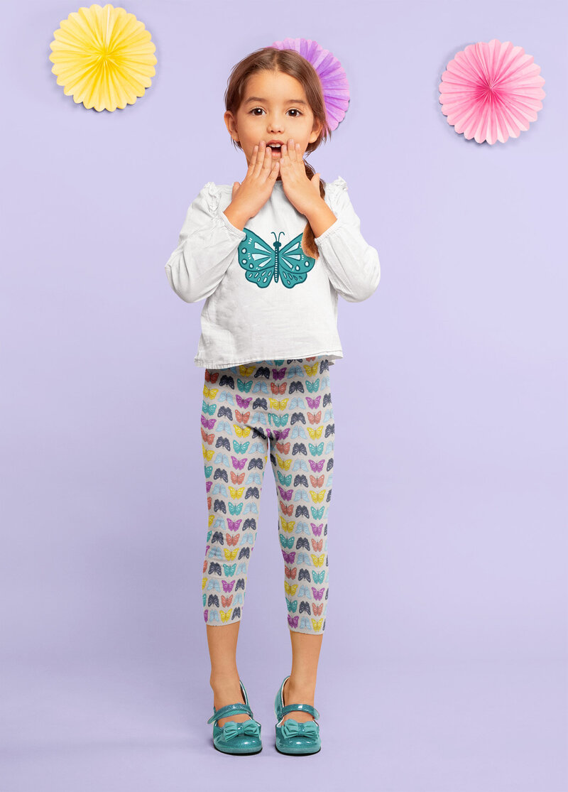 A young girl wears a butterfly themed outfit, featuring a shirt with a teal illustrated butterfly on it and all over printed leggings with butterflies in pink, yellow, orange, teal, blue and purple. Butterfly artwork by Skye McNeill.