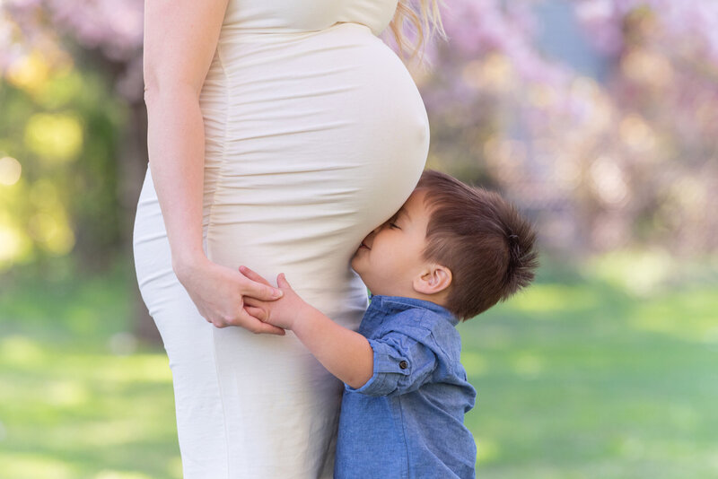 A boy kissing his mom's pregnant belly at a park.
