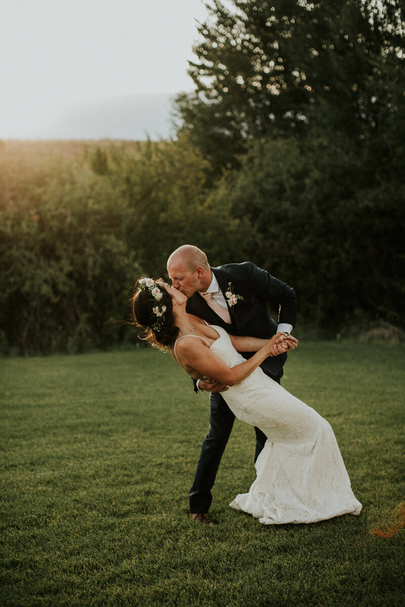 A groom dips his wife as her kisses her in a grassy yard