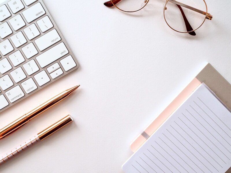 white desk with white keyboard, notebooks, rose gold pens and glasses