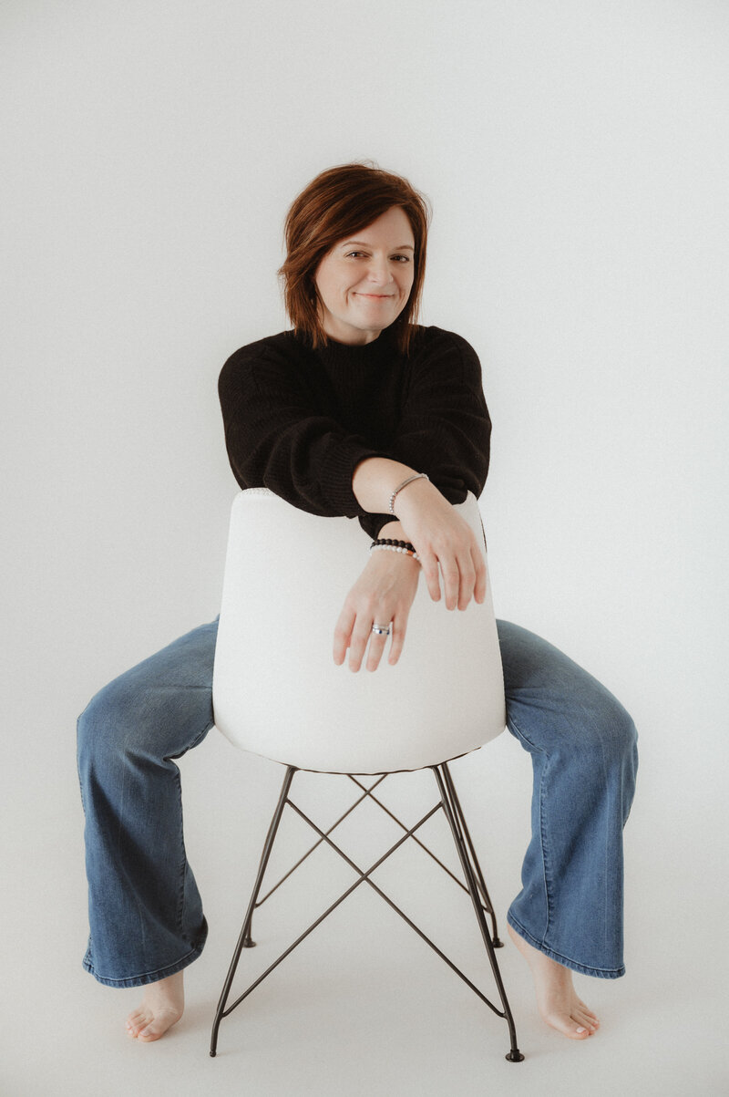 A person in a black sweater and jeans sits backward on a white chair, gazing at the camera with the distinct style of a Macon family photographer, set against a plain backdrop.