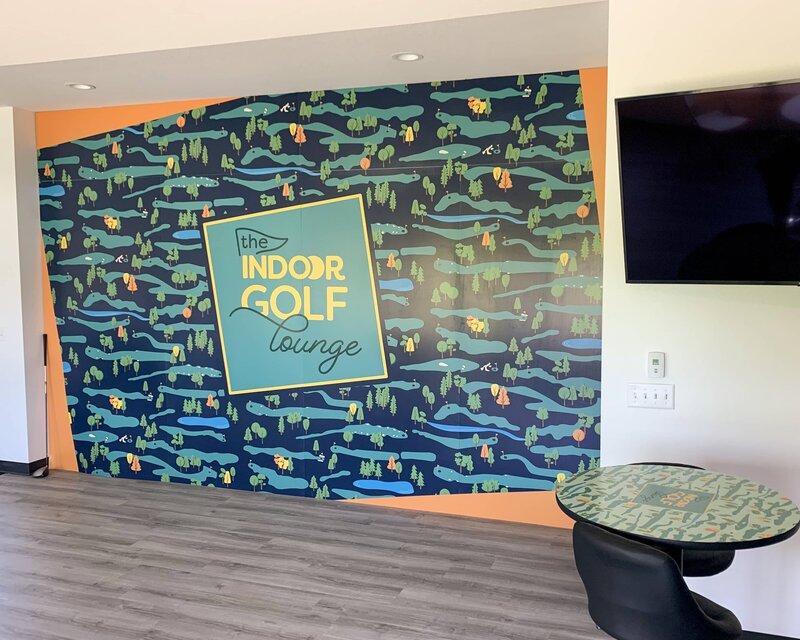 A large, interior wall covered with The Indoor Golf Lounge brand pattern with their logo in the center of the wall. A table with the brand pattern printed on top sits in one corner.