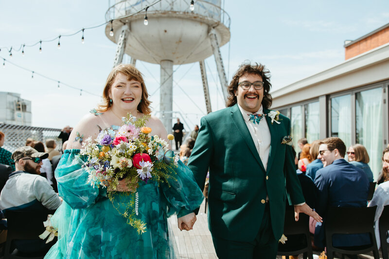 Joyful couple exiting their wedding ceremony on the Fordson Hotel rooftop in downtown Oklahoma City during a vibrant brunch celebration, with colorful floral details and a unique teal gown