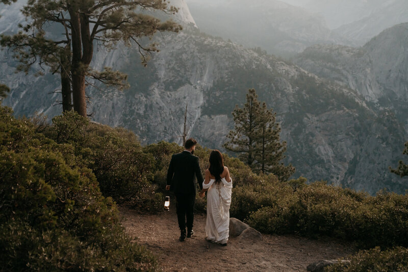 Bride and groom walking away from the camera holding hands overlooking the granite walls of yosemite valley from above, surrounded by greenery and trees