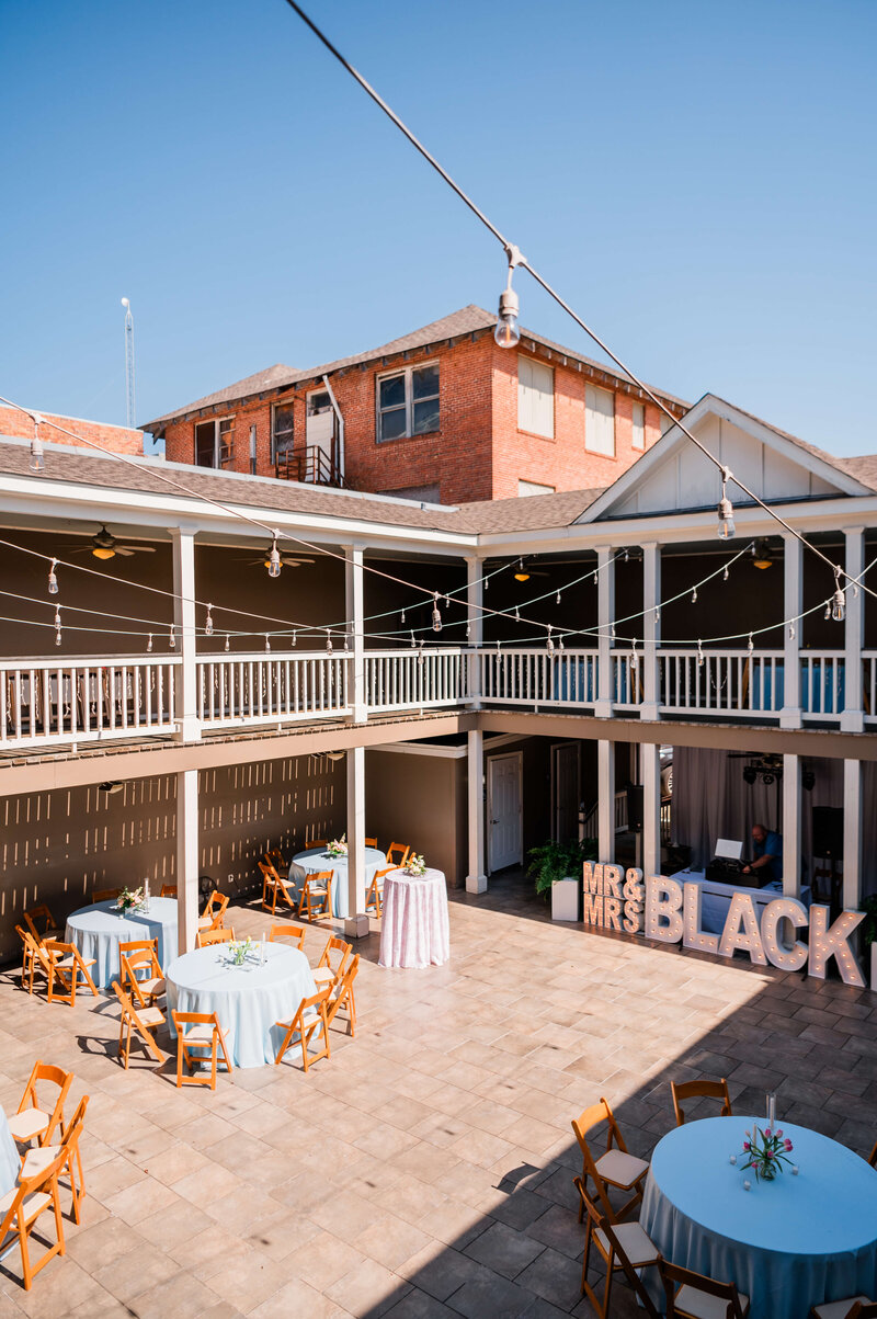 An outdoor courtyard set up for an event with round tables covered in light blue cloths, wooden chairs, string lights overhead, and large MR and MRS letters near a wall.