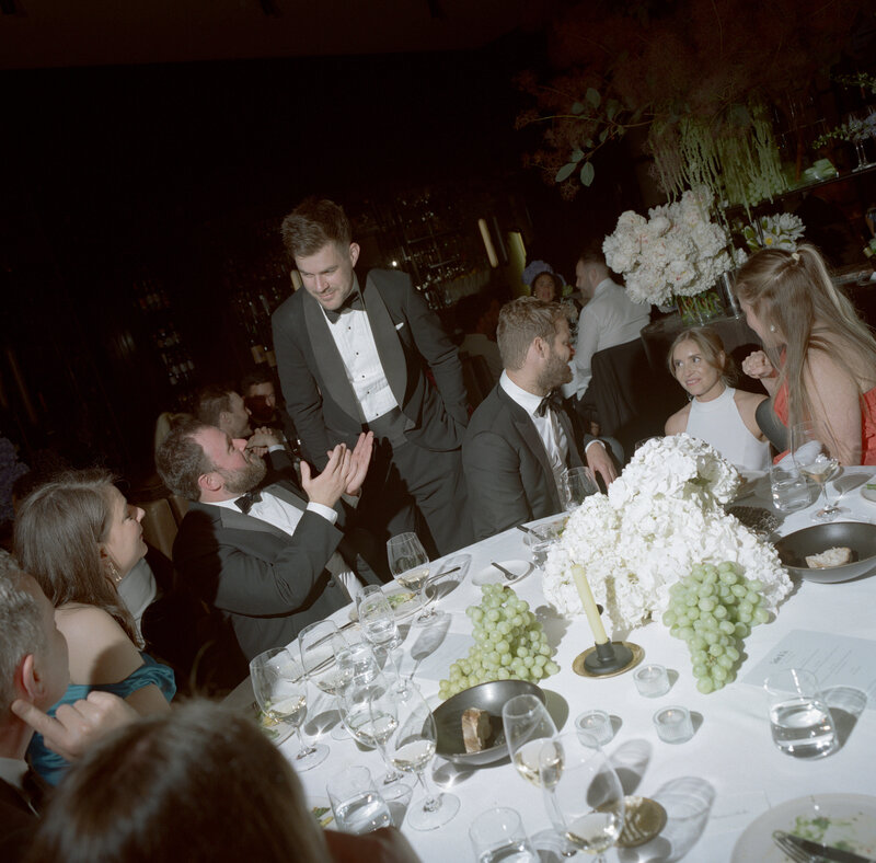 A lively, intimate wedding reception set in a dimly lit, elegant venue, Cutler and Co in Fitzroy. The scene captures a close-knit group of well-dressed guests gathered around a round table adorned with white linens, hydrangea floral arrangements, and bowls of green grapes. Most of the guests are wearing black-tie attire, adding a formal touch to the occasion. In the center, a man in a sharp tuxedo stands, leaning slightly toward another guest seated beside him, engaged in animated conversation. Glasses of white wine and water fill the table, adding to the celebratory atmosphere. The warm lighting creates soft shadows, emphasizing the relaxed yet sophisticated mood of the evening. Lush greenery and more florals can be seen in the background, enhancing the elegance of the setting.