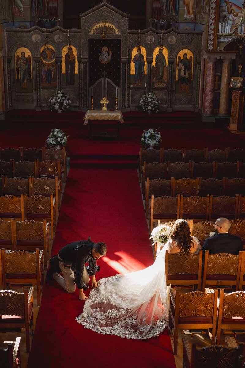 A bride and groom sit in the front row of a sunlit church as a film wedding photographer kneels on the red carpet, adjusting the bride’s long, lacy train. Rows of wooden pews and ornate icons are visible in the background.