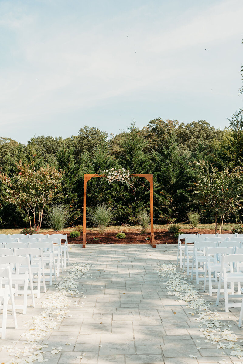 An outdoor wedding setup in Leesburg, VA, showcases a wooden arch adorned with flowers at the aisle's end. White chairs line the stone path sprinkled with rose petals. Lush trees stand under a clear sky—a perfect addition to any rustic wedding venue photo gallery.