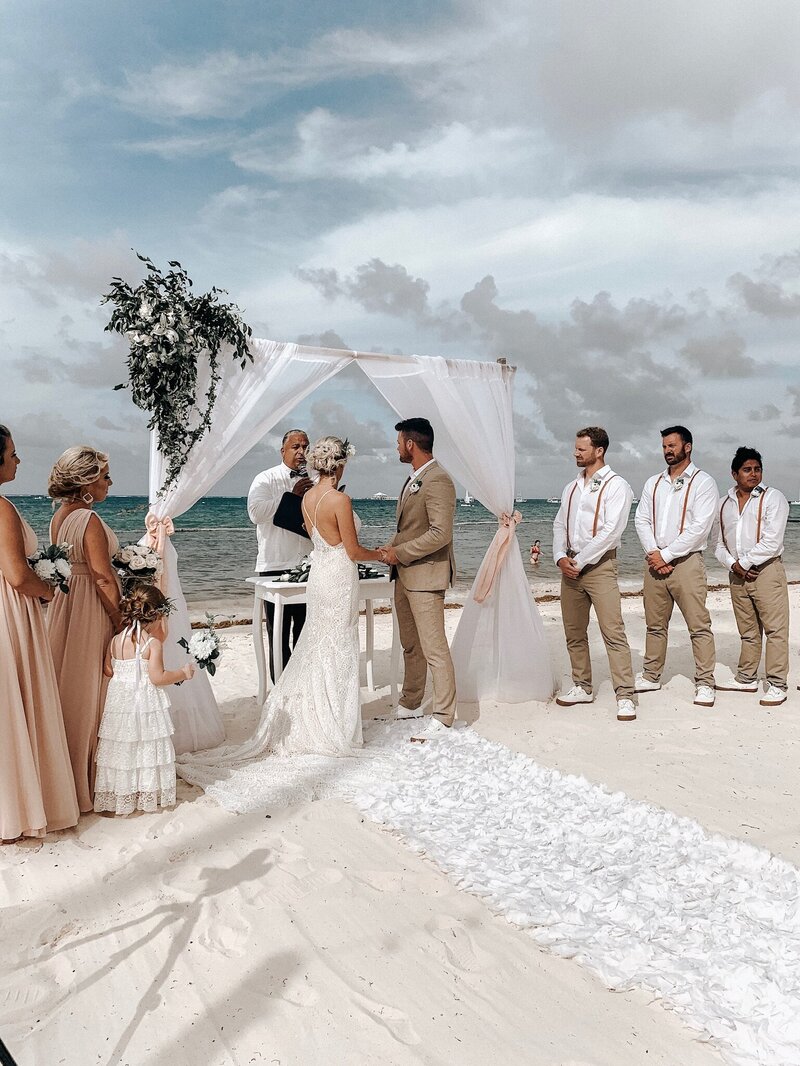 A beach wedding ceremony with the bride and groom standing under a simple white arch, exchanging vows. The bride wears a lace gown, and the groom is dressed in a tan suit. Bridesmaids in blush dresses and groomsmen in white shirts and suspenders stand alongside them, with the ocean in the background.