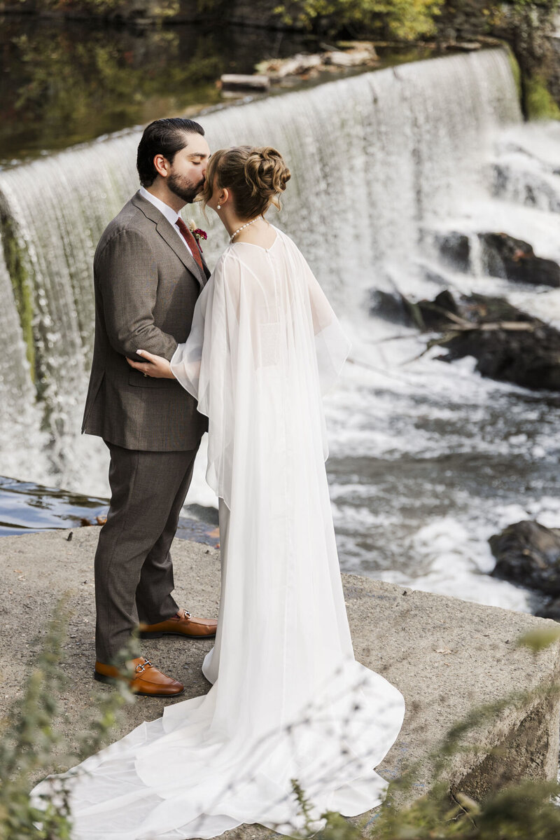 Hudson Valley wedding portrait of a couple kissing beside a waterfall with the bride in a long flowing cape veil