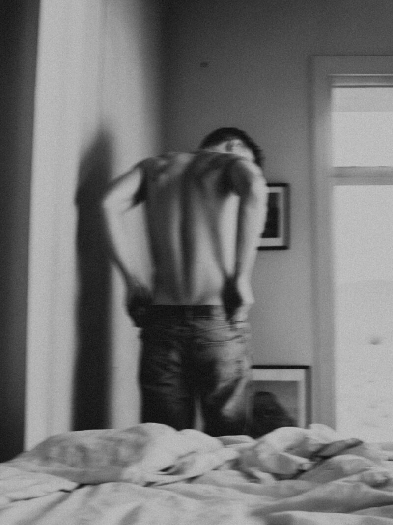 A softly lit black and white photograph of a shirtless man pulling on his jeans, his back turned to the camera. The bed behind him remains unmade, while framed artwork lines the wall, adding depth to the scene.