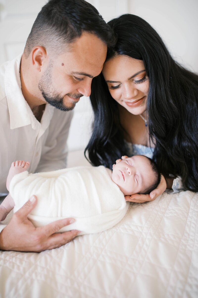 Newborn portrait taken in home in Jacksonville with mom wearing a blue patterned dress as her and dad both hold their second baby boy and look down at him taking in his details