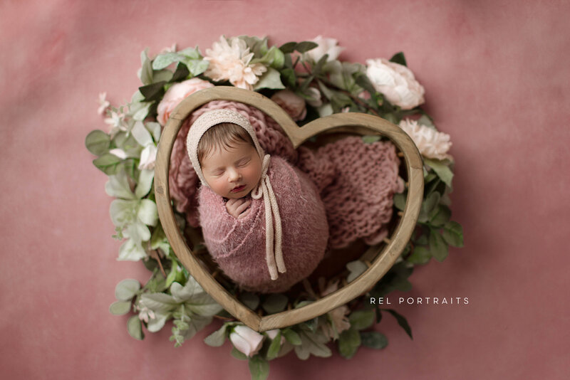 Newborn baby in Chicago swaddled in a soft pink blanket, peacefully sleeping in a heart-shaped wooden bowl surrounded by delicate greenery and flowers against a soft pink background in chicago