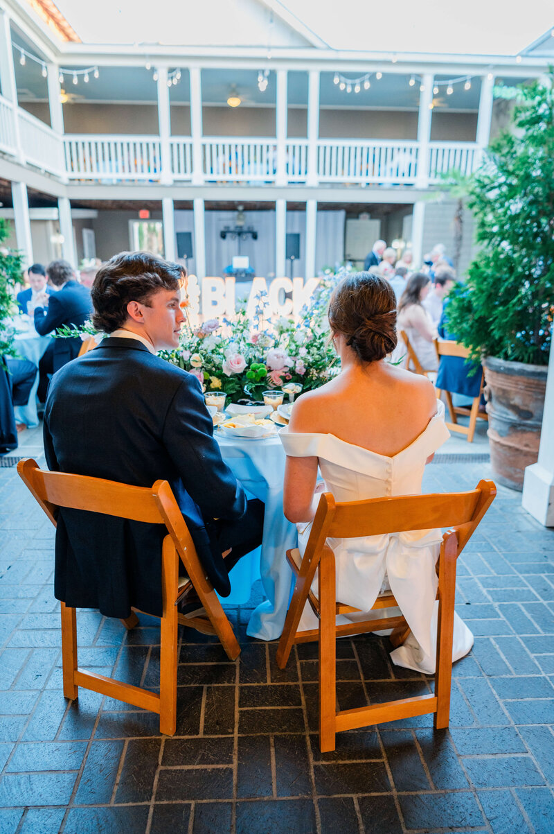 A bride and groom sit side by side at a decorated table during their wedding reception in a courtyard, facing a floral arrangement and illuminated sign, with guests seated at nearby tables.