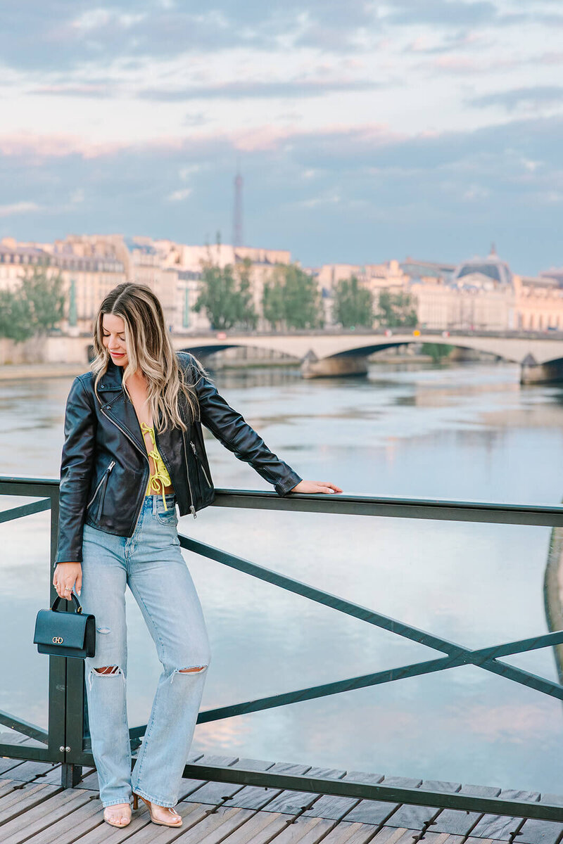 Stylish solo woman in a leather jacket and jeans, smiling confidently during a Paris photoshoot near the Louvre and Pont des Arts.