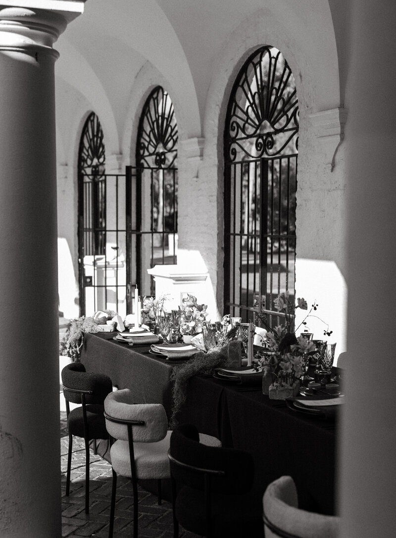 Elegant outdoor wedding table setup under arched colonnade with wrought iron gates in soft black and white light