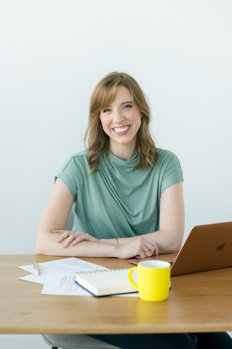 Kelsey Kemp smiling and sitting at a table with a notebook, laptop, and yellow mug