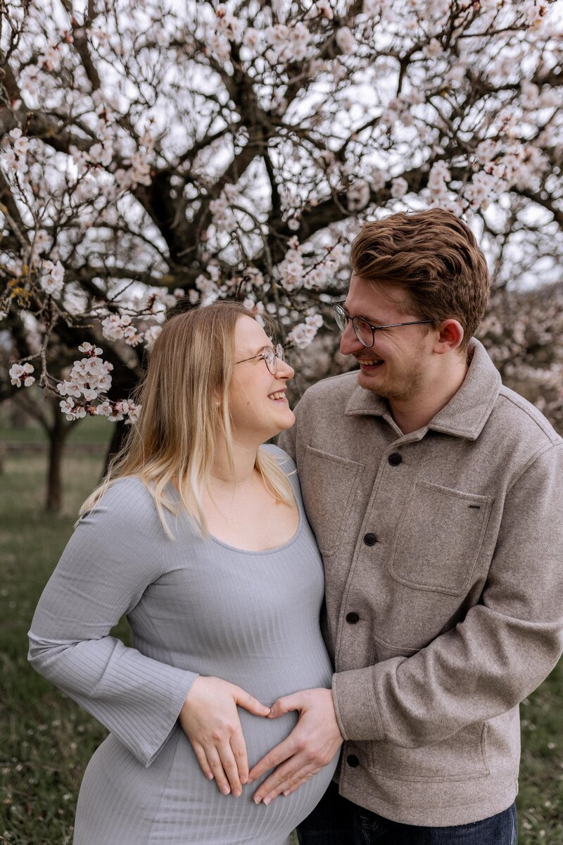 Paar steht vor einem Baum mit Kirschblüten und hält verliebt den Babybauch der Frau