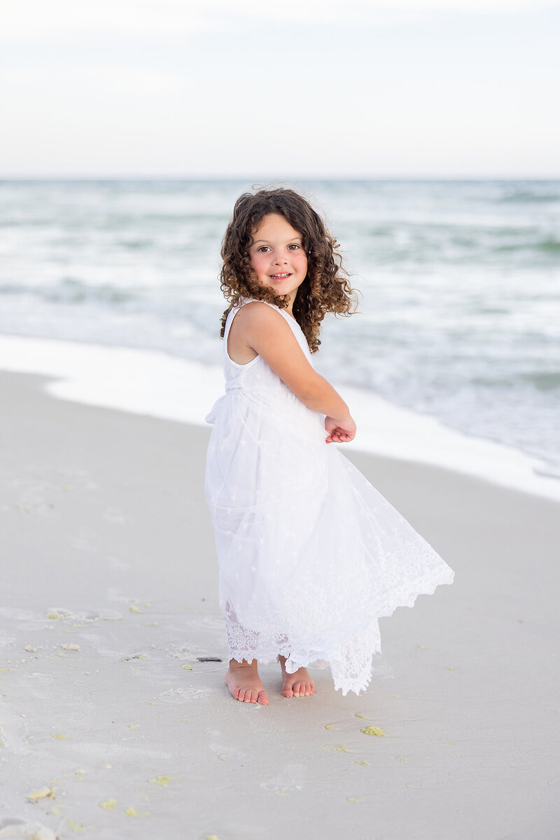 Smiling happy little girl twirling around on the beach at the waters edge at Alys Beach, Florida.  https://gwynegrayphotography.com
