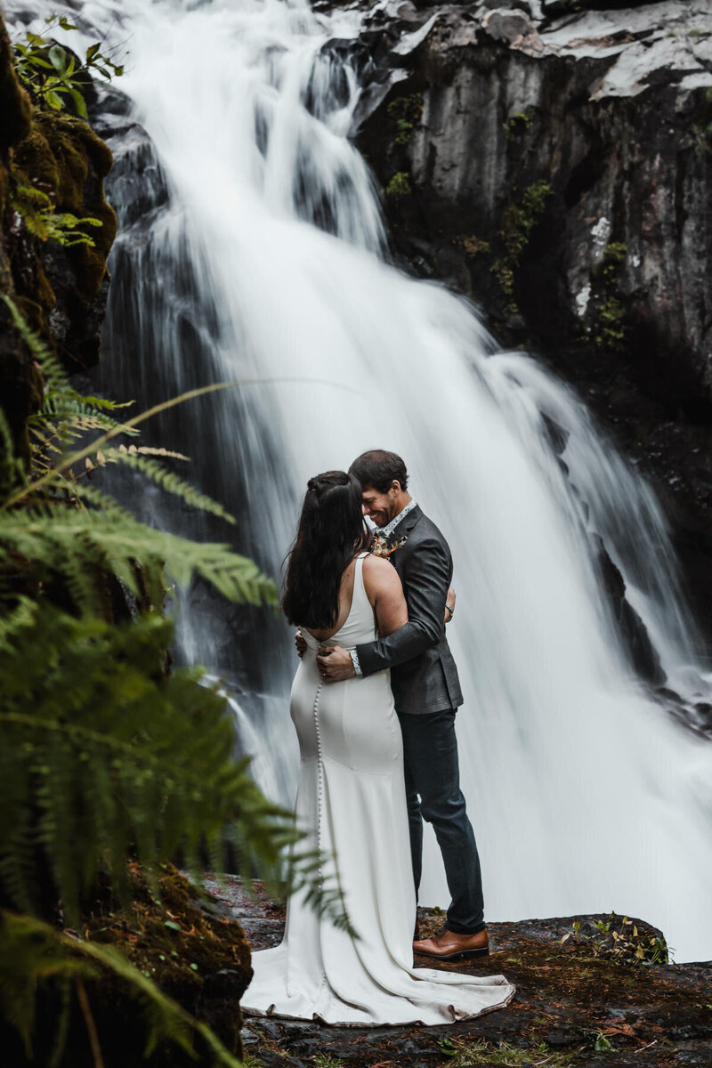 after learning how to elope in Washington State, a couple embraces in their wedding attire, as a waterfall  gushes behind them