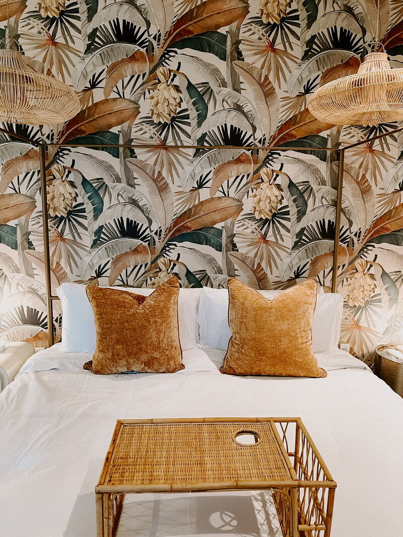 white bed with orange throw pillows against wallpaper of palm leaves