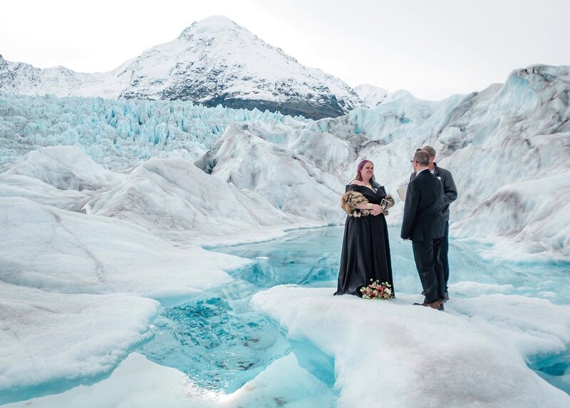 Glacier elopements in Alaska by a blue pool on a glacier--this one is next to the couple saying their vows is reflecting the snow capped mountain visible behind them.   The bride is wearing a long black wedding dress.  There is a wall of white glacier ice off to the right of them and a craggy glacier ice fall of blue and white behind them.  