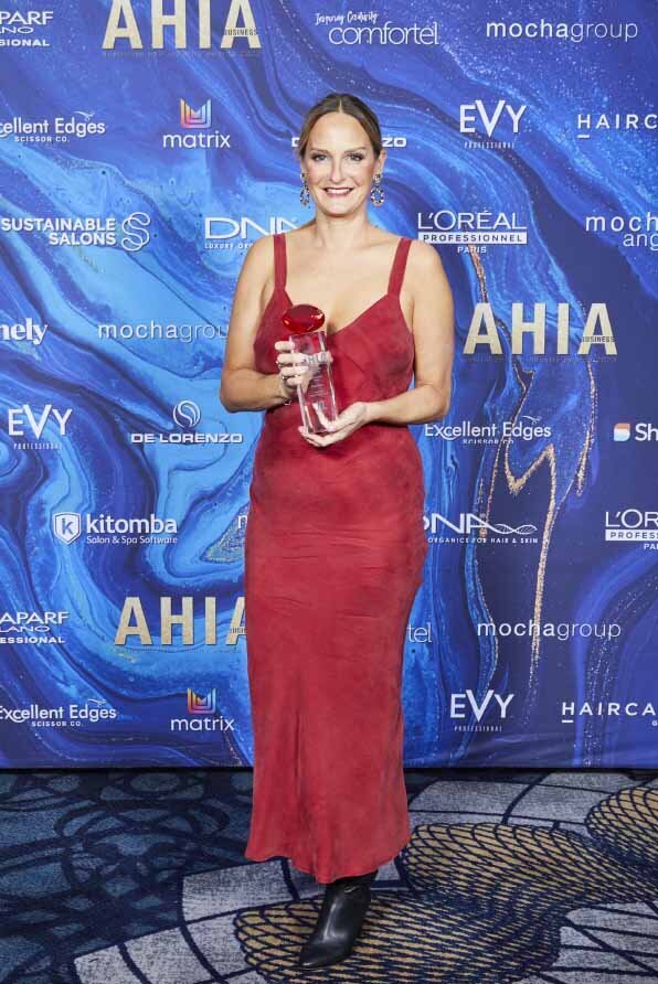 Katy Reeve in a red dress holding an award at the Australian Hair Fashion Awards, standing in front of a branded blue backdrop.